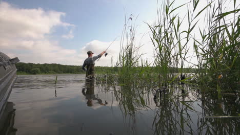 Fisherman-in-a-wader-suit-standing-in-the-water-an-trowing-a-fishing-rot-with-parallaxing-reeds-at-Lac-De-Madine-in-France
