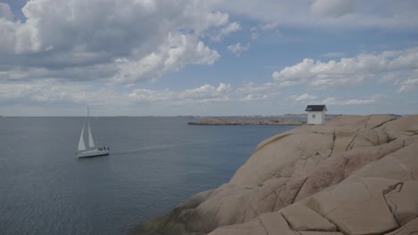 Kleines-Haus-An-Der-Küste-In-Schweden-Auf-Einem-Felsen-Mit-Vorbeifahrendem-Segelboot