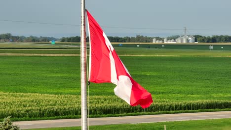 Canadian-flag-waving-with-corn-fields,-farmland,-farms,-and-grain-elevators-in-the-distance