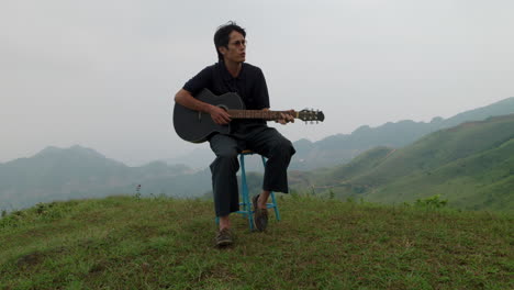 Front-view-of-young-adult-playing-black-guitar-in-middle-of-exotic-green-hilltop