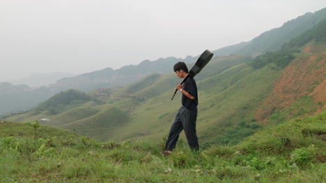 Stylish-musician-walking-with-guitar-on-shoulder-through-grassy-mountains-in-southeast-Asia