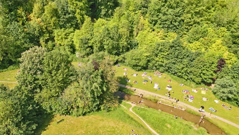 A-UK-rural-park-captured-by-drone,-showcasing-people-relishing-a-meandering-stream-and-scenic-picnic-spots,-all-within-a-woodland-covered-tourist-magnet