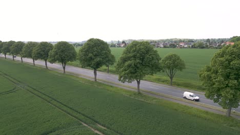 Aerial-drone-shot-of-a-road-with-cars-and-bus-passing-by,-surrounded-by-green-farmlands-along-rural-countryside-of-Brunswick,-Germany-on-a-cloudy-day