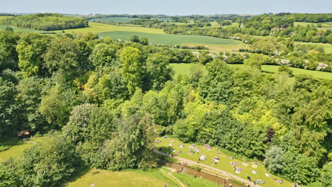 Drone-footage-captures-a-British-summer-park—individuals-enjoying-a-meandering-stream,-charming-picnic-spots,-and-a-wooded-tourist-destination
