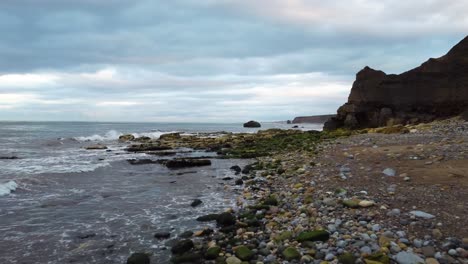 Drone-shot-flying-over-a-moss-covered-rocky-beach-on-a-cold-grey-evening,-with-an-arch-rock-formation-at-the-base-of-the-cliffs