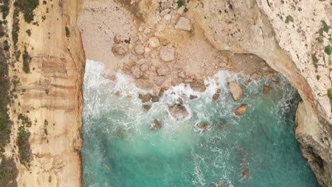 Aerial-overhead-drone-footage-of-a-hidden-vibrant-turquoise-bay-with-white-waves-crashing-on-the-beach-and-light-brown-rocks-off-Gozo's-coast,-sea-spray-soaring