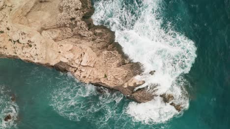 Overhead-drone-footage-reveals-stunning-turquoise-waters-as-white-waves-crash-on-a-bright-brown-rock-off-Gozo's-coast,-sea-spray-soaring-high