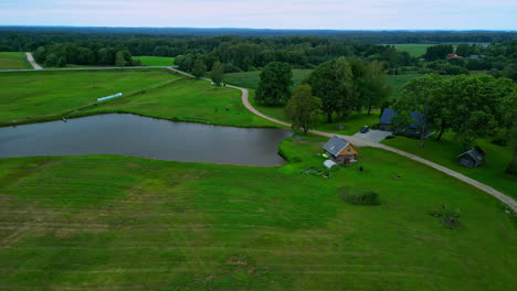 Aerial-View-of-a-Lake-with-Wooden-House-Surrounded-by-Grass-Fields-and-Woodland-in-Latvia