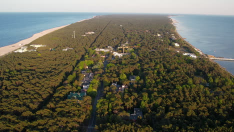 Aerial-backwards-shot-over-green-forest-on-Hel-Island-and-working-cranes-at-construction-site-in-Jurata-Village-at-sunny-day---beautiful-landscape-with-Baltic-sea-and-sandy-beach-in-poland