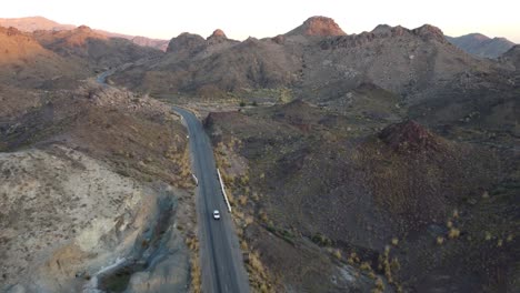 Drone-shot-revealing-a-car-driving-on-road-carved-through-mountains-at-sunset,-Balochistan,-Pakistan