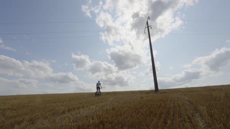 Man-rides-mountain-bike-through-open-grassy-farm-fields-below-concrete-utility-pole,-blue-sky