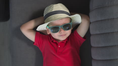 Süßes-Kleinkind-Mit-Sonnenbrille-Und-Hut-Liegt-Lächelnd-Auf-Der-Couch