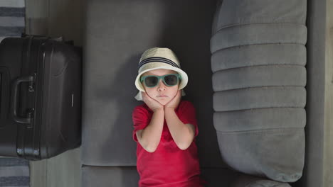 Adorable-toddler-adjusts-straw-hat-lying-on-soft-grey-sofa