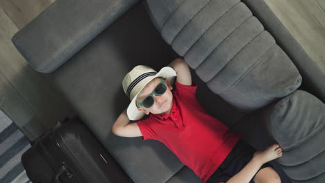 Adorable-child-in-summer-hat-relaxes-on-couch-near-suitcase