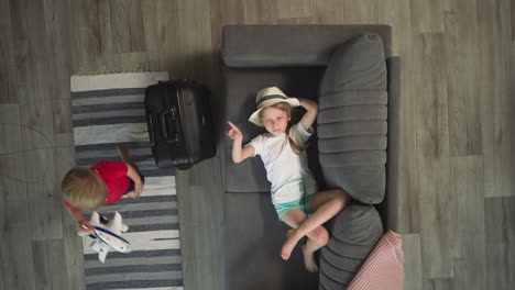 Toddler-walks-past-sister-resting-on-sofa-and-big-suitcase
