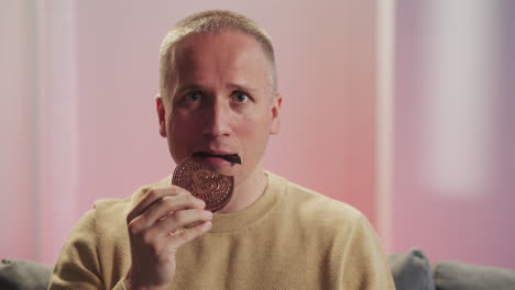 Blond-man-bites-piece-of-chocolate-cookie-looking-in-camera