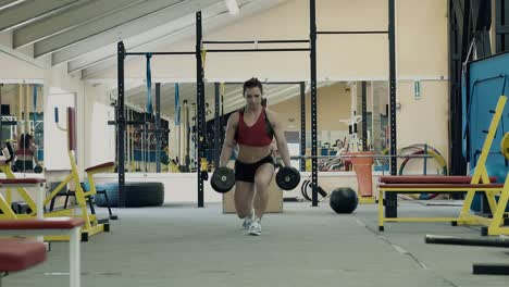 Una-Joven-Atlética-En-El-Gimnasio-Pasa-Por-El-Pasillo-Agachada-Con-Pesas