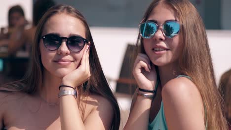 Dos-Jóvenes-Atractivas-Chicas-Calientes-Con-Gafas-De-Sol-Están-Sentadas-A-La-Mesa-En-La-Playa
