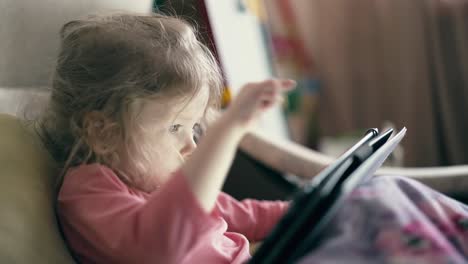 CU-portrait-of-pretty-little-girl-child-who-lies-on-sofa-under-blanket-and-plays-on-tablet-computer