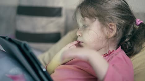 CU-portrait-of-pretty-little-girl-child-who-lies-on-the-sofa-under-blanket-and-plays-on-a-tablet-computer