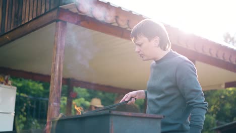 young-man-standing-near-the-summer-house-prepares-the-coals-in-the-grill-fanning-them