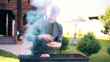 Young-man-stands-on-the-background-of-a-summer-house-lays-marinated-pork-on-a-barbecue-grill