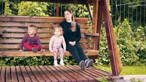 Travelling-Young-pretty-mother-with-children-swinging-on-a-wooden-swing-near-the-summer-house-2