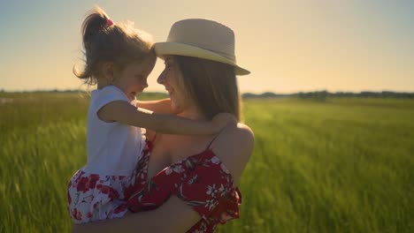 Close-up-Young-mother-wearing-hat-holds-little-daughter-in-her-arms-standing-in-field-the-wind-blows-hair-flutters