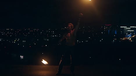 Young-blond-male-spins-two-burning-torches-on-night-city-background-Slow-motion