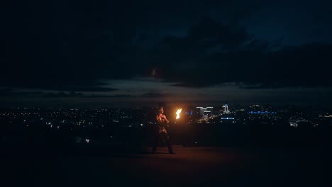 Young-blond-male-does-fire-show-in-the-middle-of-the-night-with-city-skyline-in-background-spins-burning-torch