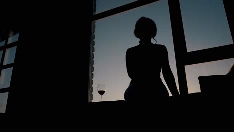 Silhouette-Einer-Schlanken-Dame,-Die-Neben-Einem-Weinglas-Auf-Der-Fensterbank-Sitzt