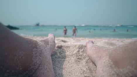 young-man-feet-on-sandy-beach-against-blue-ocean-with-people