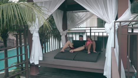 couple-with-devices-rests-on-hotel-wooden-terrace-at-pool