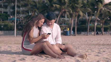 barefoot-couple-sits-on-sand-and-girl-types-on-smartphone