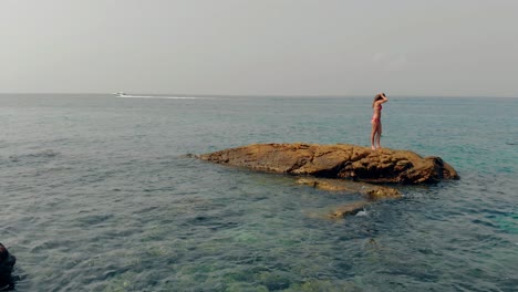 girl-stands-on-stone-midst-clear-blue-water-against-boat