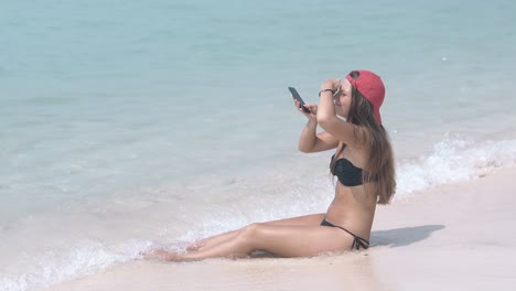 long-dark-haired-woman-sits-on-beach-water-edge-with-phone
