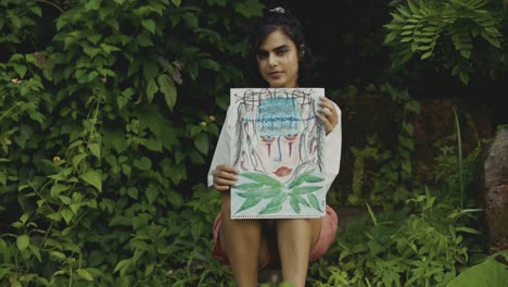 An-beautiful-girl-sitting-between-plants-showing-a-drawing-with-an-engaging-stare
