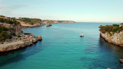 Catamaran-Boat-In-Crystal-Clear-Water-Overlooking-The-Es-Pontas-In-Cala-Llombards,-Mallorca,-Spain