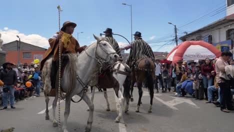 Erleben-Sie-Einen-Kulturellen-Parade-Moment,-Wenn-Zwei-Reiter,-Die-Elegant-Auf-Pferden-Sitzen-Und-Ponchos-Und-Hüte-Tragen,-Unter-Der-Führung-Einer-Acocha-Autorität-Die-Straße-Entlangschreiten