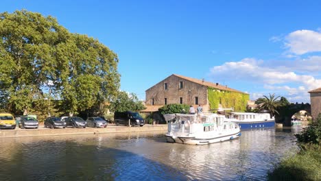 Canal-Du-Midi-En-Le-Somail-Francia-Barco-Turístico-Pasando-Por-El-Pueblo-Y-Bajo-El-Puente-En-Una-Cálida-Tarde-De-Verano