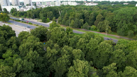 Aerial-backwards-shot-of-green-forest-and-driving-cars-on-intersection-in-Warsaw-City,Poland