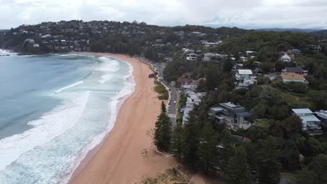 Drone-flying-over-a-beachside-road-revealing-expensive-real-estate-on-the-cliffs-and-a-beautiful-sandy-beach