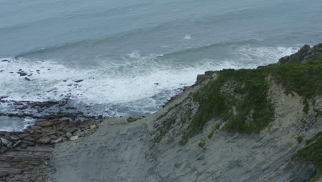 Drone-dolly-tilt-up-reveals-crashing-waves-on-rocky-slabs,-itzurun-beach