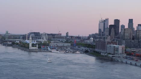 Aerial-view-of-a-Sailboat-in-the-St-Lawrence-River-at-Sunset-with-the-Montreal-Skyline,-Port-of-Montreal,-and-the-Ferris-Wheel-in-the-Background