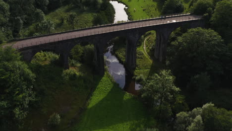 Aerial-view-tilting-over-a-bridge,-revealing-cattle-grazing-on-meadows-at-a-river-in-Peak-district,-UK