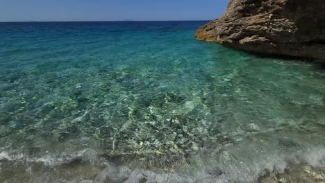 Crystal-emerald-water-reflecting-sunlight-on-seabed-with-pebbles-sheltered-by-cliffs-on-Mediterranean-shoreline