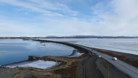 Aerial-view-following-cars-on-a-bridge-at-a-frozen-lake,-sunny,-winter-day-in-Iceland