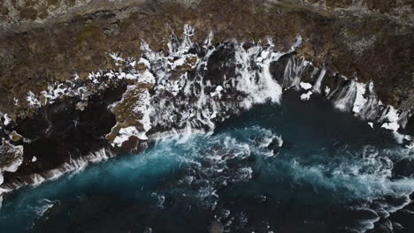 Aerial-view-rising-away-from-the-snowy-Hraunfossar-waterfall,-in-cloudy-Iceland