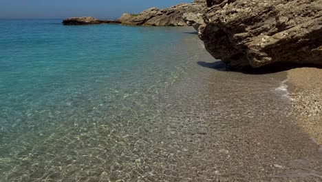 Big-cliffs-splashed-by-calm-seawater-on-beautiful-quiet-beach-with-pebbles-and-crystal-emerald-water-in-Mediterranean