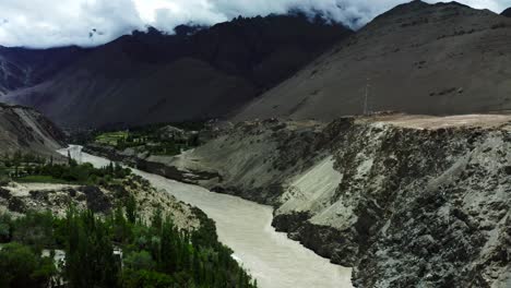 Aerial-drone-camera-moving-forward-in-Leh-Ladakh-mountain-village-with-river-flowing-in-stream-and-clouds-moving-over-mountain